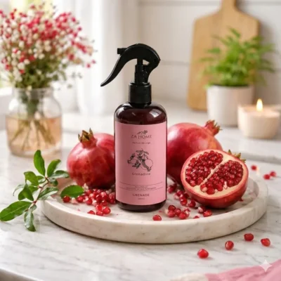 Spray parfum de linge ZA Home senteur Grenadine 250ml, posé sur un plateau en marbre avec des grenades entières et une coupée, et loose arils. Formule clean sans PEG ni huile de ricin hydrogénée. Ambiance fruitée et gourmande pour la maison en Tunisie.